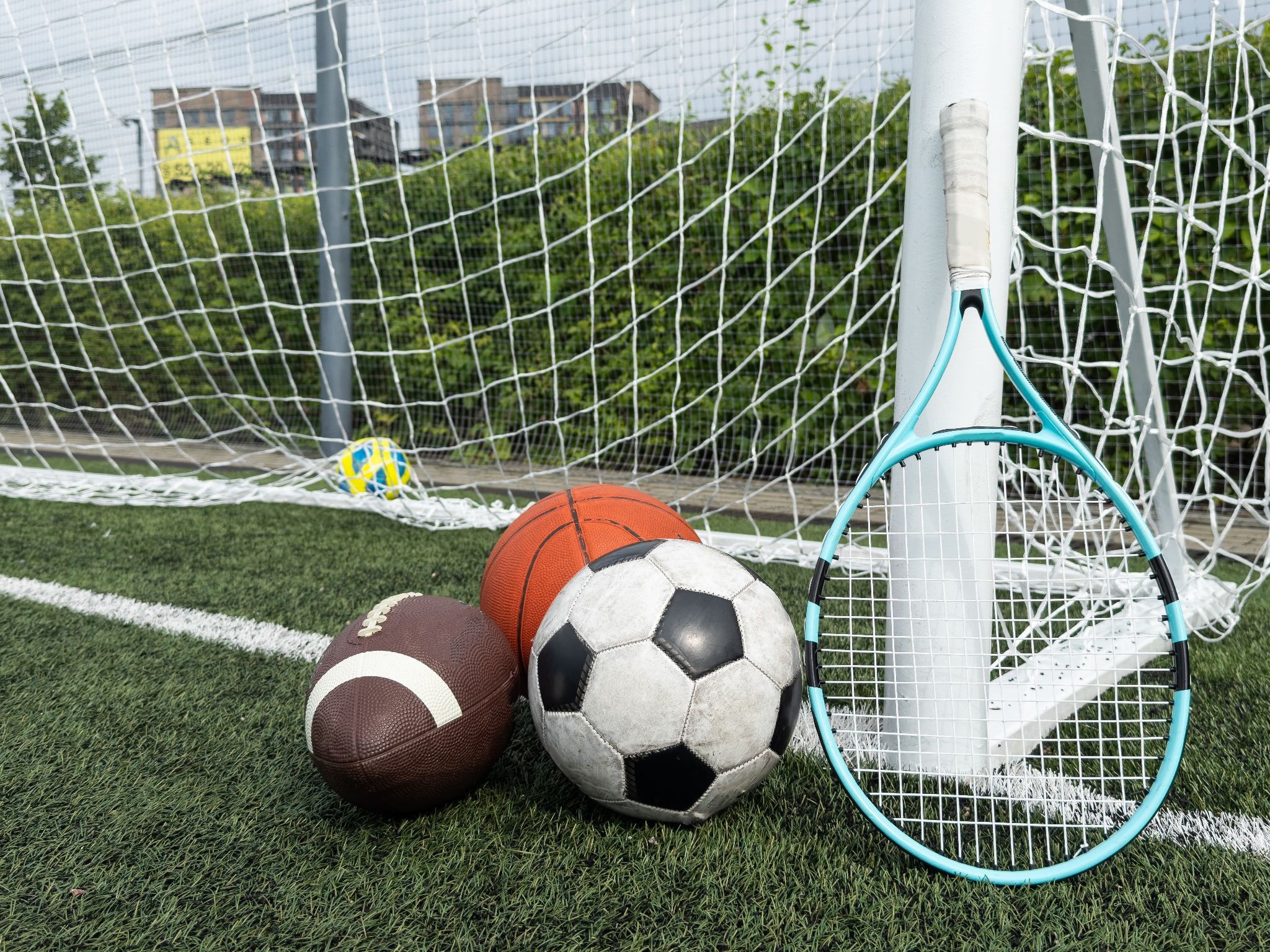 różne piłki i rakieta tenisowa przy bramce piłkarskiej z siatką ochronną w tle